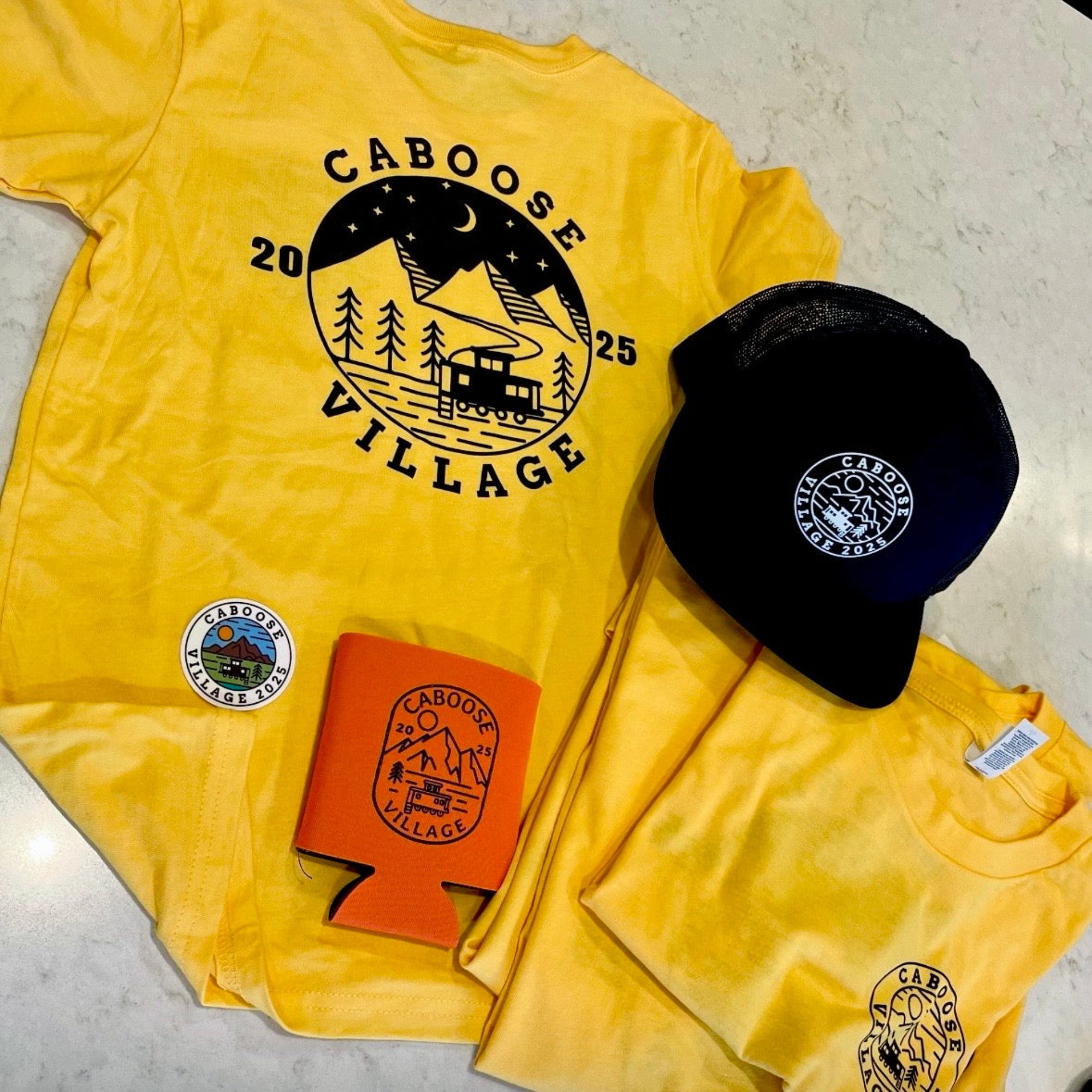 Yellow t-shirt with 'Caboose Village' design, black cap, and orange can cooler on a light gray surface.